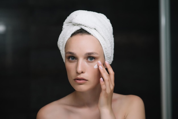 Photo gros plan portrait de tête de belle femme appliquant une crème hydratante sur le visage après la douche jeune jolie femme enveloppée dans une serviette lissant perfectionnant le concept de routine quotidienne du matin