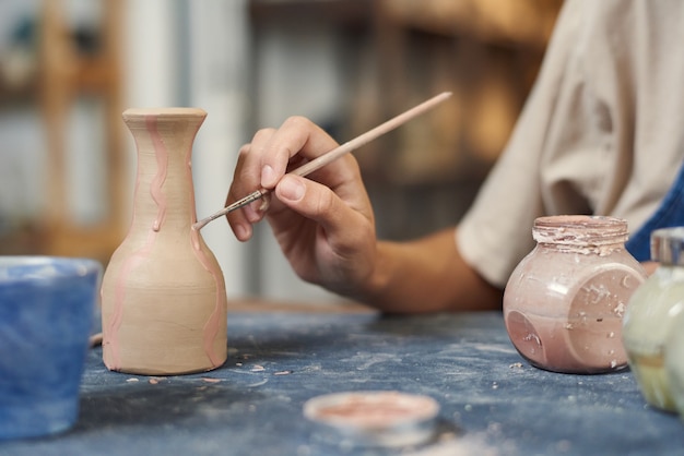 Gros plan d'un potier méconnaissable à l'aide d'un pinceau tout en appliquant de la peinture rose sur un vase d'argile en atelier