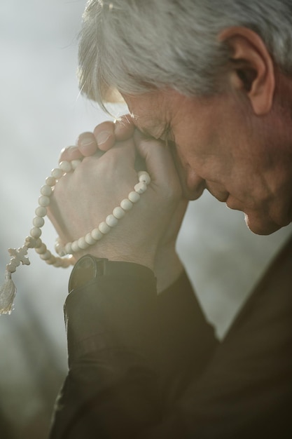 Gros plan d'un prêtre senior aux cheveux blancs priant avec un chapelet et un éclairage serein