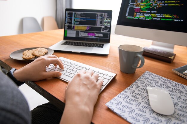 Gros plan d'un programmeur méconnaissable assis au bureau avec du café et des cookies et création de code