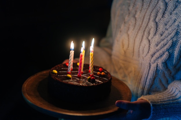 Gros plan recadré d'une jeune femme méconnaissable tenant dans les mains un gâteau d'anniversaire avec six bougies allumées dans une pièce sombre