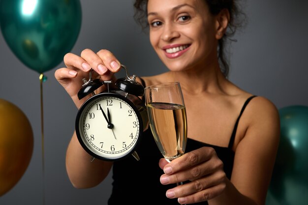 Gros plan d'un réveil et d'une flûte de champagne dans les mains d'une belle femme souriante floue, isolée sur fond gris et ballons à air métalliques verts et dorés gonflés, espace de copie pour l'annonce