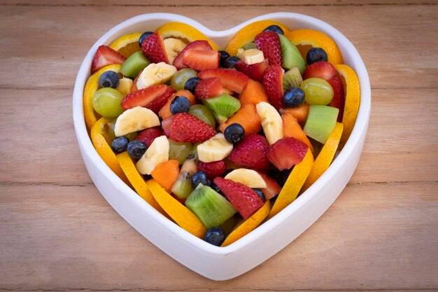 Gros plan d'une salade de fruits colorés mélangés dans une assiette en céramique blanche en forme de coeur. Fond en bois