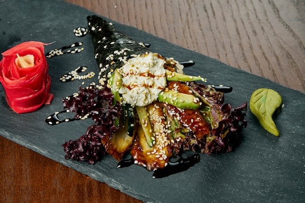 Gros plan de savoureux sushis roulés à la main dans des mamenori avec de l'anguille et du caviar Tobico servis sur une plaque en pierre sombre avec de la sauce soja et du gingembre .. Temaki, cuisine japonaise. Alimentation sainea