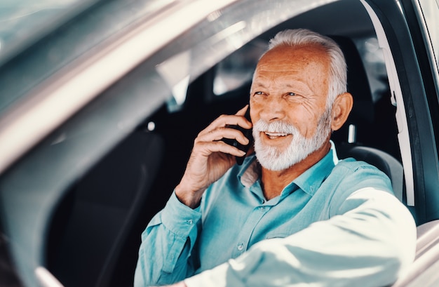 Gros plan de senior barbu souriant parlant au téléphone avec le bras sur la fenêtre ouverte tout en étant assis dans la voiture.