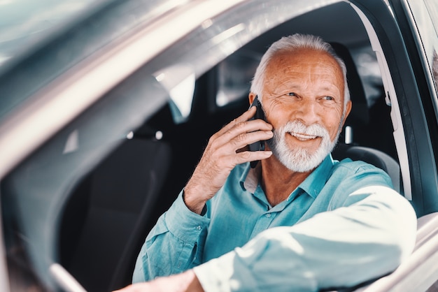 Gros plan de senior barbu souriant parlant au téléphone avec le bras sur la fenêtre ouverte tout en étant assis dans la voiture.