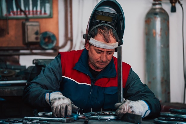 Gros plan soudeur en uniforme de protection et masque de soudage de tuyaux métalliques sur la table industrielle avec d'autres travailleurs derrière dans l'atelier industriel