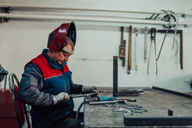 Gros plan soudeur en uniforme de protection et masque de soudage de tuyaux métalliques sur la table industrielle avec d'autres travailleurs derrière dans l'atelier industriel