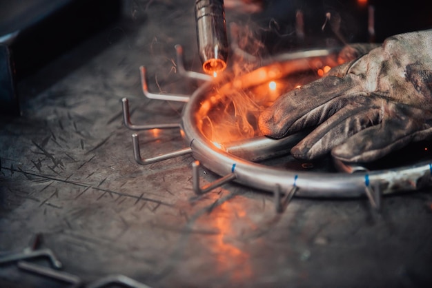 Gros plan soudeur en uniforme de protection et masque de soudage de tuyaux métalliques sur la table industrielle avec d'autres travailleurs derrière dans l'atelier industriel