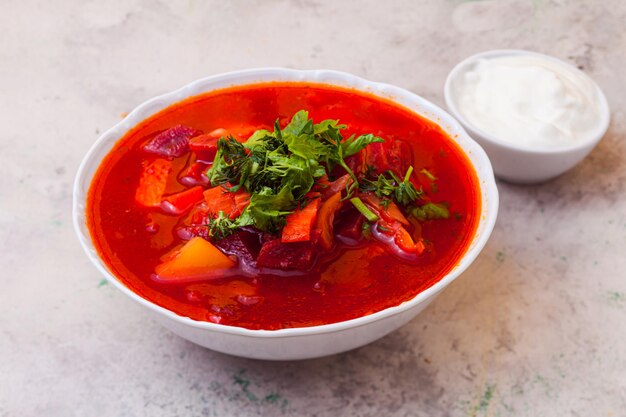 Gros plan de soupe traditionnelle ukrainienne et russe avec des légumes verts et de la crème sure à côté de la soupe de betterave dans une assiette sur le tableau blanc