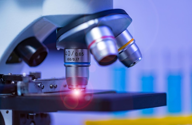 Gros plan en studio d'une caméra de microscope à lentille de zoom à loupe placée sur une table de travail dans un laboratoire hospitalier plein d'équipement de verrerie support de tube à essai bécher de fiole Erlenmeyer et écran d'ordinateur.