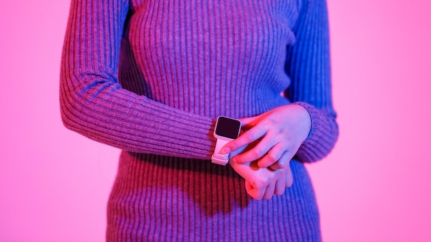 Gros plan en studio d'un gadget à la mode de smartwatch à écran tactile vide numérique noir vierge sur le poignet de la femme qui en robe pull grise à manches longues pointant avec l'index sur fond rose clair.