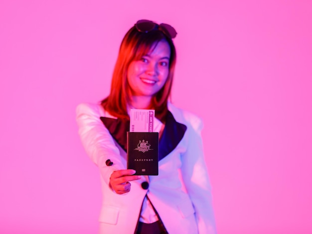 Gros plan en studio d'un passeport de voyage et d'un billet d'avion à la main d'une voyageuse riche et urbaine asiatique en costume de blazer décontracté et des nuances regardent la caméra sur fond rose clair flou.