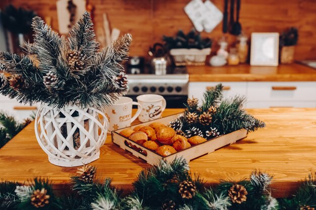 Gros plan de la table de cuisine avec vase de branches de sapin, deux tasses en céramique et plateau en bois avec des petits gâteaux faits maison et des pommes de pin avec des branches de sapin.