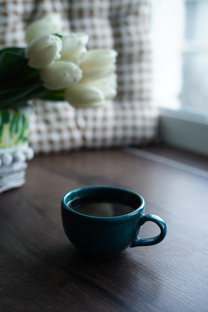 Un gros plan d'une tasse de café noir turquoise contre un panier de tulipes blanches. Une belle tasse bleue