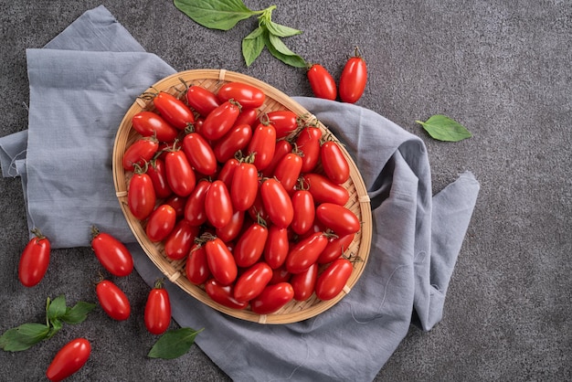 Gros plan de tomates cerises fraîches dans un panier avec feuille de basilic sur fond de table gris.