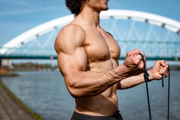 Gros plan sur un torse nu d'un bodybuilder fort avec six packs, un ABS parfait, des épaules, des biceps, des triceps et de la poitrine, qui fait un entraînement corporel solide avec un élastique, près de la rivière.