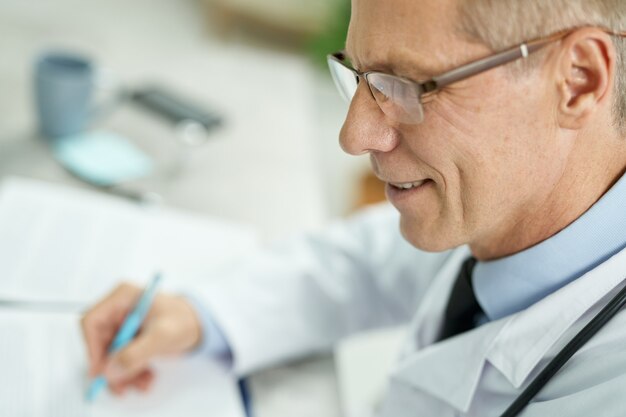 Gros plan d'un travailleur médical joyeux tenant un stylo et souriant tout en étant assis à la table au travail