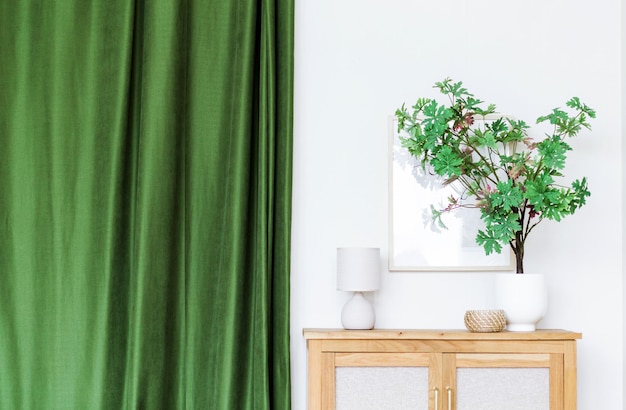Gros plan sur un vase élégant avec une plante verte sur un placard, une lampe et un rideau vert