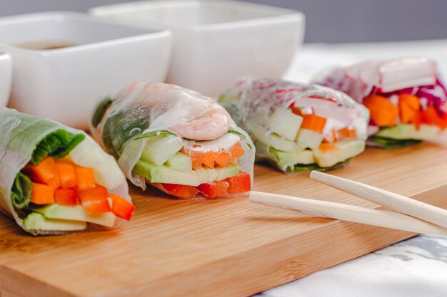 Gros plan vertical de rouleaux de printemps frais avec des crevettes et des légumes coupés en deux et différentes sauces servies sur une plaque de bois.