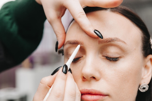 Gros plan sur le visage d'une fille Le maître tient un crayon blanc et dessine la forme des sourcils pour le maquillage permanent