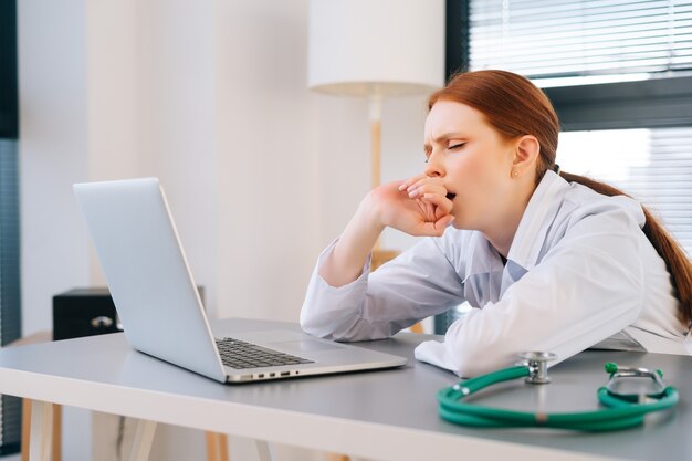 Gros plan sur le visage d'une jeune femme médecin fatiguée et épuisée portant un manteau blanc qui bâille pendant qu'elle travaille sur un ordinateur portable.