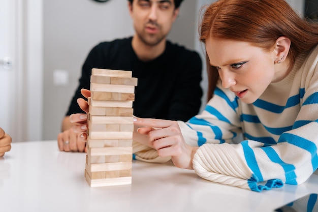 Gros plan sur le visage d'une jeune femme rousse sérieuse et concentrée retirant soigneusement le bloc de bois de la structure de la tour en bois Diverses personnes concentrées assises à table