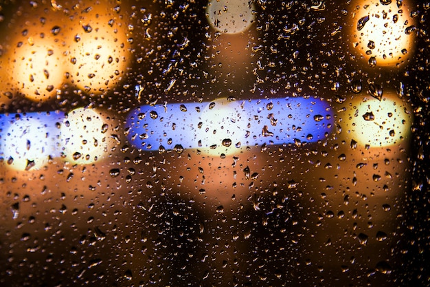 Gros plan sur une vitre de voiture avec une touffe de pluie, en arrière-plan une lueur jaune vif