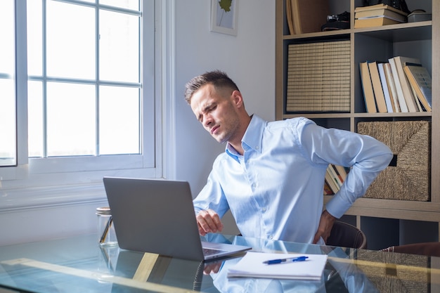 Gros plan sur la vue arrière du jeune homme stressé touchant le bas du dos ressentant une gêne, souffrant d'une douleur soudaine due à un mode de vie sédentaire ou à un long surmenage informatique dans une posture incorrecte au bureau à domicile.