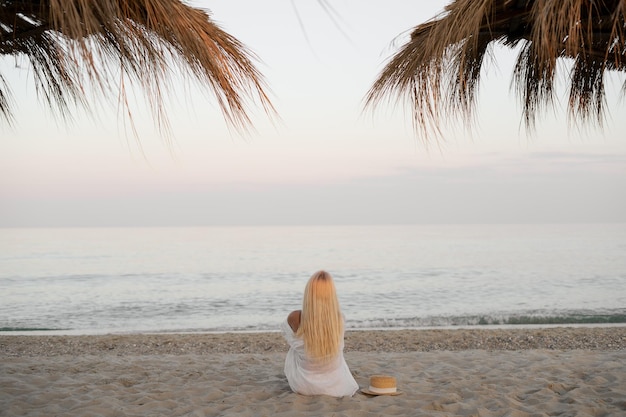 Gros plan vue arrière d'une femme assise dans une robe blanche sur le sable au bord de l'océan et regardant au loin à la surface de l'eau un chapeau de paille se trouve à proximité Mouvement de la caméra