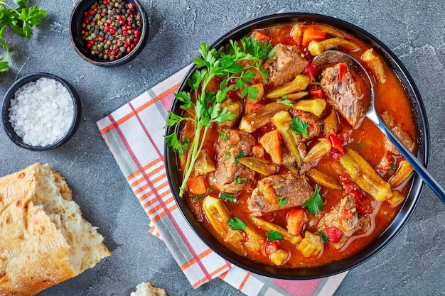 Gros plan de la vue de dessus de délicieux ragoût de boeuf et de gombo servi dans un bol avec cuillère, sur une table en béton gris avec du pain ciabatta, mise à plat