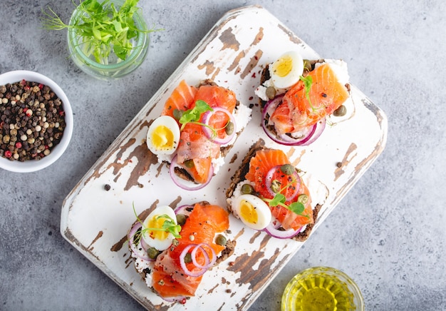 Gros plan, vue de dessus du sandwich avec pain de seigle noir, fromage à la crème, saumon, oignon, câpres, œuf à la coque, planche à découper en bois blanc, fond en béton. Sandwich ouvert traditionnel scandinave Smorrebrod