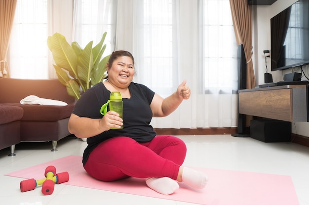 Grosse femme asiatique tenant une bouteille d'eau avec le pouce levé faisant de l'exercice à la maison, concept d'idée de sport et de loisirs.