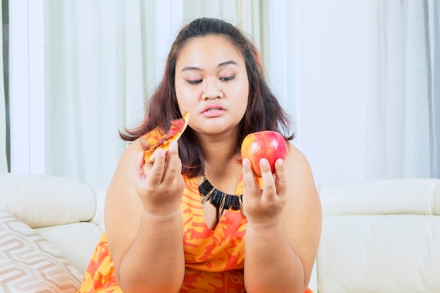 Une grosse femme confuse choisit une pomme ou une pizza sur un canapé