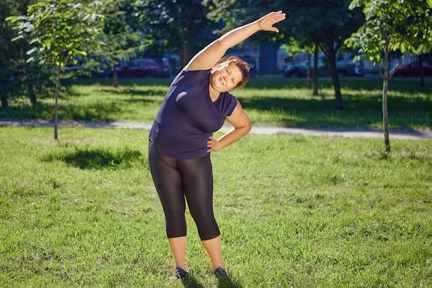 Grosse femme européenne fait des exercices de virages latéraux debout sur la pelouse dans le parc