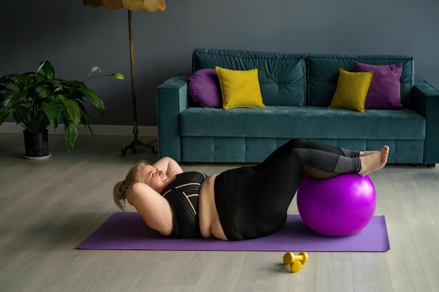 Une grosse femme potelée fait de l'exercice pour soulever le tronc à la maison en mettant ses pieds sur une balle en forme