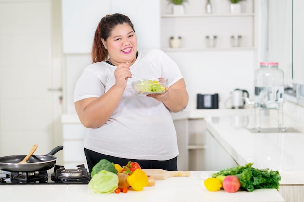 Une grosse femme souriante mange une salade saine dans la cuisine.