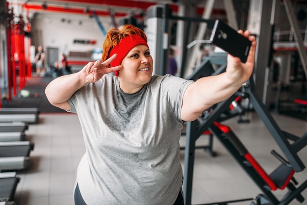 Grosse femme en sueur fait selfie contre les machines d'exercice dans la salle de gym. Calories brûlées, personne de sexe féminin obèse dans un club de sport