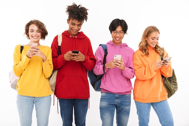 Groupe d'adolescents joyeux isolés, portant des sacs à dos, à l'aide de téléphones mobiles
