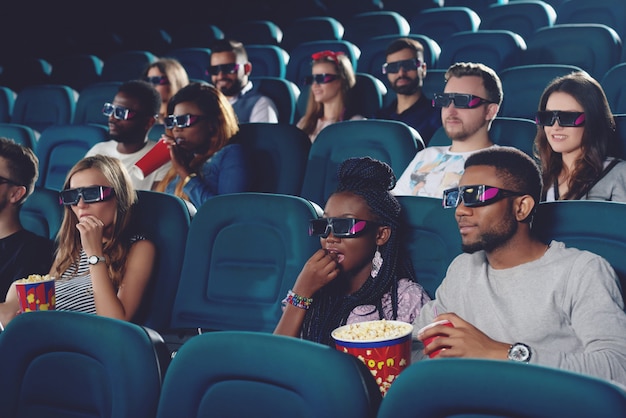 Groupe d'africains et de caucasiens, regarder un film dans des lunettes 3d dans une salle de cinéma moderne.