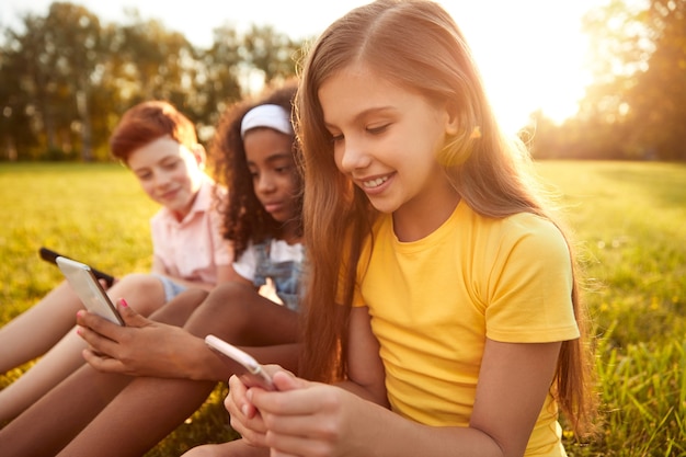 Groupe d'amis d'âge scolaire heureux assis sur l'herbe et utilisant des smartphones tout en passant la journée d'été ensemble dans la nature