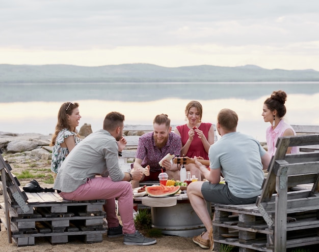 Groupe d'amis assis autour d'une table près du lac pittoresque le soir d'été, bavardant, mangeant et profitant de leur temps ensemble