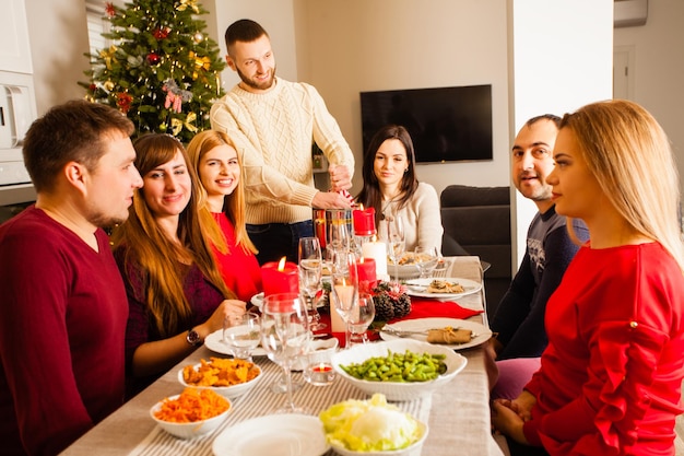 Photo groupe d'amis assis sur la table à la veille de noël. sélibérer ensemble la fête du nouvel an, la nourriture et les boissons. l'homme ouvre le champagne