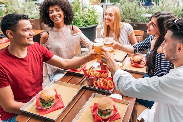 Groupe d'amis gais multiethniques se réunissant à table avec de la restauration rapide et des verres de bière trinquant tout en proposant des toasts et en célébrant l'anniversaire d'un ami