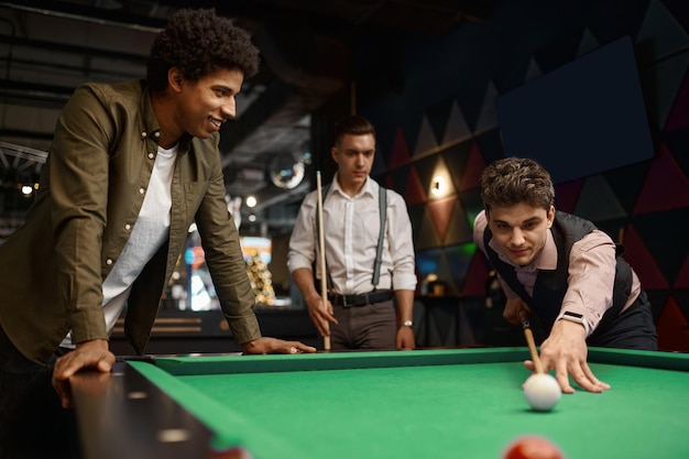 Groupe d'amis heureux appréciant le jeu de billard, compagnie de beaux jeunes gars jouant dans la piscine