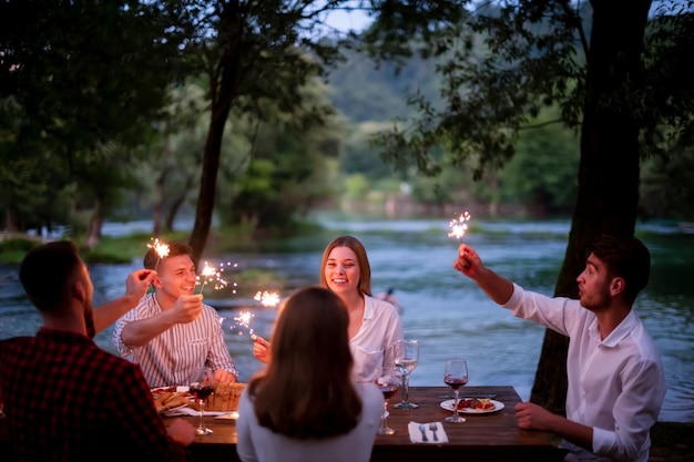 groupe d'amis heureux célébrant des vacances de vacances en utilisant des arroseurs et en buvant du vin rouge tout en pique-niquant un dîner français en plein air près de la rivière lors d'une belle soirée d'été dans la nature