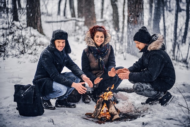 Un groupe d'amis heureux a organisé un camping au milieu d'une forêt enneigée, assis autour d'un feu de camp et se réchauffant les mains