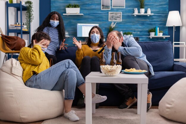 Groupe d'amis multiethniques regardant un film d'horreur à la télévision ayant peur, passant le week-end ensemble portant un masque facial pour prévenir l'infection par le covid 19 pendant la pandémie mondiale en mangeant de la bière et des frites