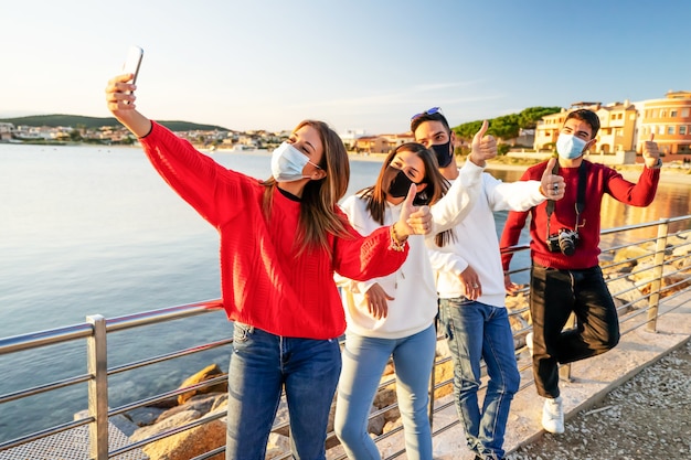 Groupe d'amis portant un masque de protection du visage contre le coronavirus faisant un selfie en ligne montrant les pouces vers le haut