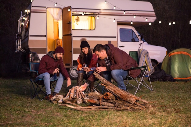 Groupe d'amis proches dégustant une bière ensemble et faisant un feu de camp avec leur camping-car rétro en arrière-plan.